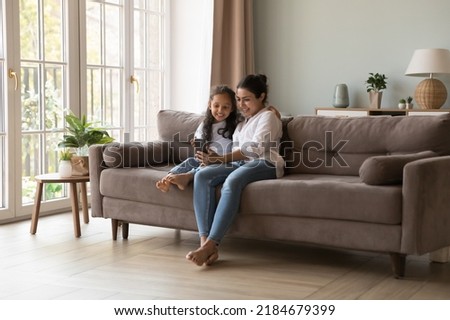 Happy Indian mom and kid taking home selfie on mobile phone, resting on sofa in living room interior, laughing, hugging, holding smartphone in outstretched hand, making video call. Full length Royalty-Free Stock Photo #2184679399