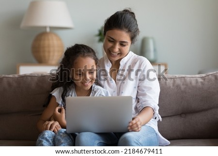 Happy pretty Indian mother and cheerful cute girl kid sharing laptop, enjoying leisure on couch together, watching movie, talking on video call, looking at monitor, smiling, laughing, hugging Royalty-Free Stock Photo #2184679411