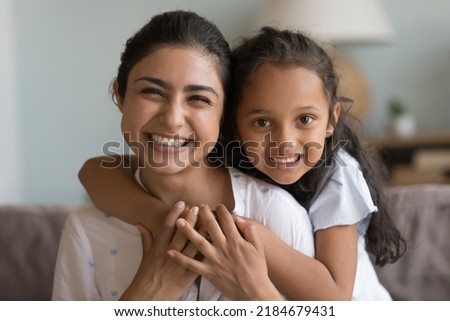 Happy beautiful young Indian mom and cute daughter kid home head shot. Cheerful mother piggybacking child looking at camera with toothy smile, hugging, talking on video call. Screen view portrait