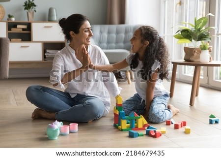 Happy beautiful Indian mom and pretty little daughter girl clapping hands, giving high five over wooden construction blocks, toy tower, castle on heating floor, celebrating model completing