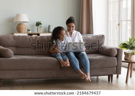 Happy pretty Indian mother showing learning app on laptop to little daughter. Mom and cute girl watching movie together, talking on video call, enjoying leisure in home living room cozy interior