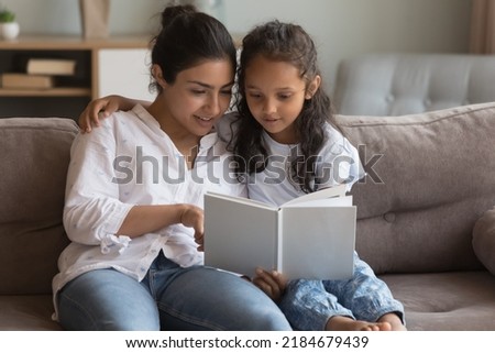 Happy young Indian mom teaching little daughter to read book, helping with primary school homework, showing pictures, telling fairy tale. Mother and kid girl resting on comfortable sofa