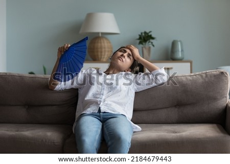 Exhausted young Indian girl feeling hot, tired, sick, resting on couch at home, waving paper handheld fan for cooking, suffering from heat attack, hypoxia, headache, touching head Royalty-Free Stock Photo #2184679443