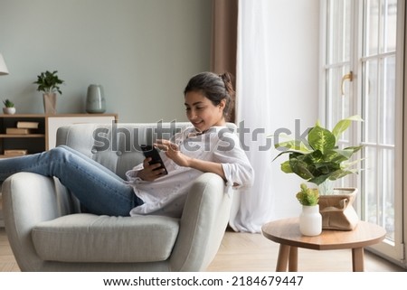 Cheerful relaxed young Indian woman using mobile phone, resting in cozy soft armchair in modern home Interior, shopping with online internet ecommerce app, touching screen, smiling