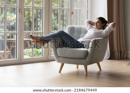 Calm peaceful lazy young Indian homeowner woman resting in armchair with closed eyes at big terrace window, breathing fresh cool air, enjoying peace, leisure, comfort, meditating at home Royalty-Free Stock Photo #2184679449