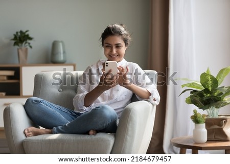 Happy pretty millennial Indian girl relaxing at home, resting in armchair, typing on smartphone, using online app, software, shopping on Internet, making video call. Mobile phone communication Royalty-Free Stock Photo #2184679457