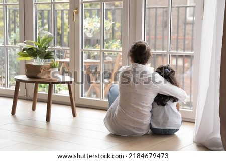 Back view of black haired young mom and little daughter girl sitting on floor at home, hugging with love, care, looking at panoramic window, cozy balcony. Motherhood, real estate concept Royalty-Free Stock Photo #2184679473