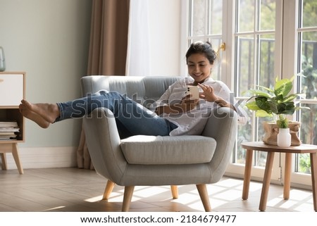 Joyful pretty millennial Indian cellphone user girl resting in armchair at cozy home, touching mobile phone screen, typing, chatting online, enjoying leisure, shopping on Internet, communication Royalty-Free Stock Photo #2184679477