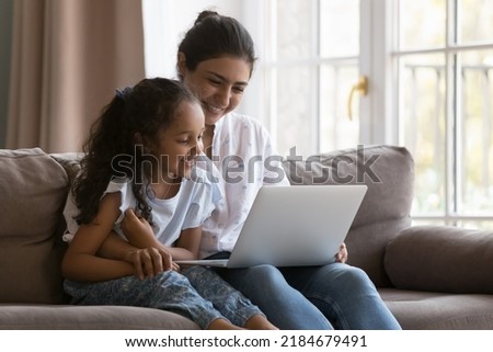 Happy young Indian mother and sweet curious kid watching cartoon movie, series, reading book online, talking on video call, using online learning app on laptop, enjoying leisure Royalty-Free Stock Photo #2184679491