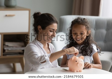 Happy excited Indian mother and kid dropping cash into piggybank. Caring mom teaching kid to save, invest money, collecting coins in piggy bank. Family savings, financial education concept Royalty-Free Stock Photo #2184679501