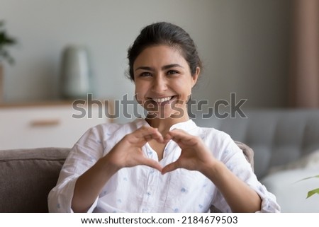 Happy pretty young Indian woman showing joined hands at chest, finger heart shape at camera, relaxing on comfortable sofa, enjoying home leisure, comfort. Blogger expressing love. Head shot portrait