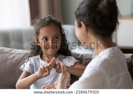 Happy cute Indian kid girl talking to mother, using hands, fingers, speaking sign languages, sitting on sofa at home. Female teacher, therapist teaching child with deafness to communicate