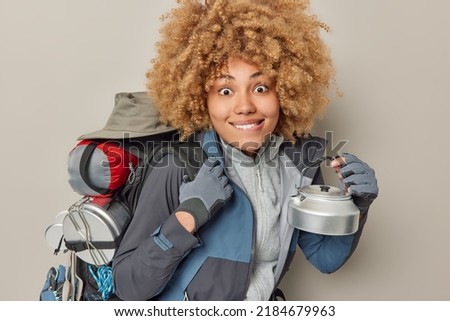 Indoor shot of curly haired woman bites lips dressed in comfortable clothes holds kettle prepares for camping and hiking looks gladfully at camera isolated over grey background. Active rest concept