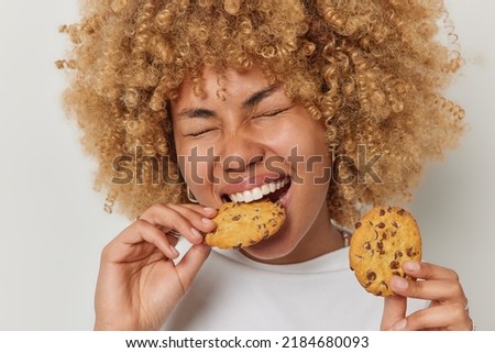 Close up shot of blonde curly haired woman eats cookies with chocolate bites deicious snack keeps eyes closed has sweet tooth dressed casually isolated over white background. Unhealthy eating Royalty-Free Stock Photo #2184680093
