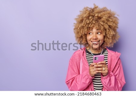 Positive curly haired young woman enjoys messaging on mobile phone dressed in casual clothes smiles happily focused away isolated over purple background with copy space for your advertisement