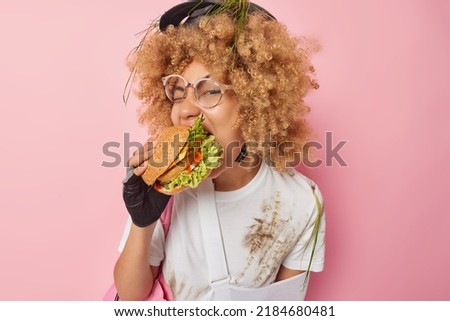 Photo of hungry young woman with blonde curly hair eats appetizing hamburger feels hungry after active rest wears helmet and white dirty t shirt isolated over pink background. Fast food concept