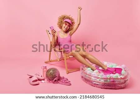 Positive curly woman celebrates good news holds mobile phone expresses happiness dressed in swimsuit poses on comfortable deck chair near swimming pool enjoys summer vacations. Perfect holiday