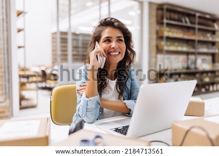 Cheerful businesswoman speaking on the phone while working in a warehouse. Happy online store owner making plans for product shipping. Female entrepreneur running an e-commerce small business. Royalty-Free Stock Photo #2184713561