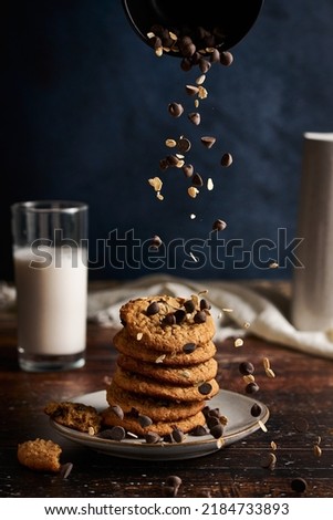 Stack of cookies on a plate. Chocolate chips and oats falling down on the dessert. Glass of Milk in the background. Wooden table. Action shot. Food Photography. High resolution. 