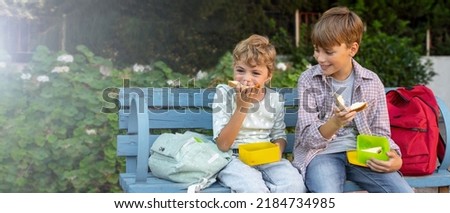 Children eating lunch sandwiches in school yard during break, sitting on the bench. Share the food, talking, laughing, resting outdoor. Back to school routine. 4K Royalty-Free Stock Photo #2184734985