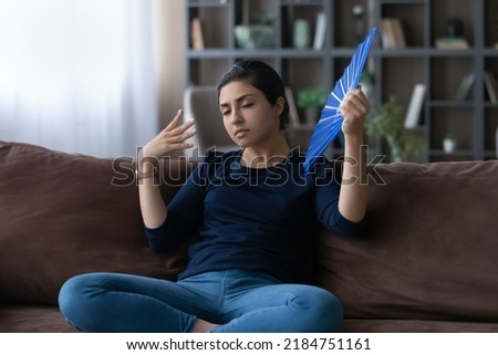 Tired millennial Indian woman sit rest on sofa at home feel unwell suffer from heatstroke wave with hand fan. Exhausted young mixed race female use waver breathe fresh air. Condition concept. Royalty-Free Stock Photo #2184751161