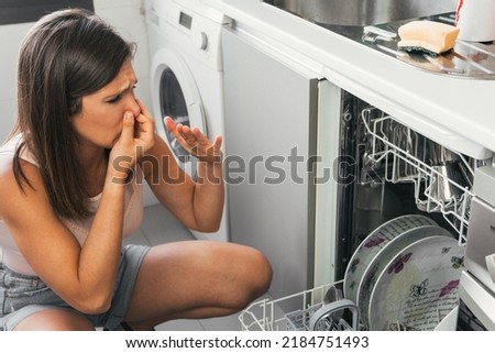 Woman making gestures of bad smell when opening her automatic dishwasher. Young girl with a face of disgust and holding her nose at the malfunction of one of her appliances. Royalty-Free Stock Photo #2184751493