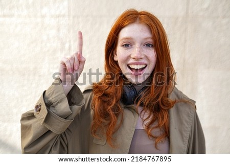 Happy teen redhead fashion girl standing on urban wall background looking at camera pointing finger up. Excited teenage girl with red hair having great new idea concept, outdoors.