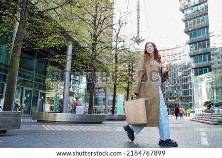 Cool fashion stylish teen girl model shopper buyer wearing trench coat holding paper shopping bags walking on busy big city street buying sale retail clothes in downtown store mall outdoors. Royalty-Free Stock Photo #2184767899