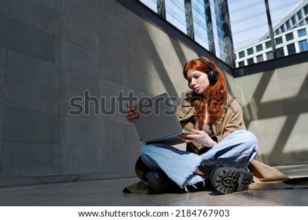 Teen redhead hipster girl student wearing headphones using laptop in city urban location searching information, online learning, watching webinar class or having video chat outdoors. Royalty-Free Stock Photo #2184767903