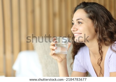 Happy woman looking away holding a water glass sitting on the bed in a bedroom 