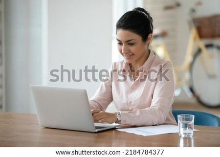 Smiling young Indian female employee sit at desk at workplace look at laptop screen consult client online. Happy mixed race woman work distant on computer with paperwork in office. Technology Royalty-Free Stock Photo #2184784377