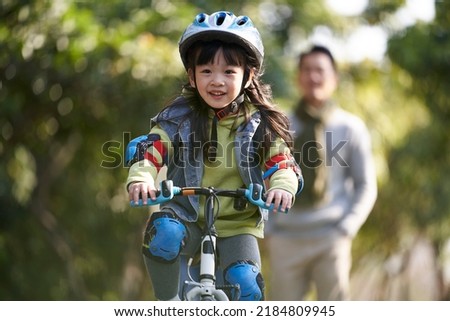 little asian girl with helmet and protection gear riding bike in city park with father watching from behind