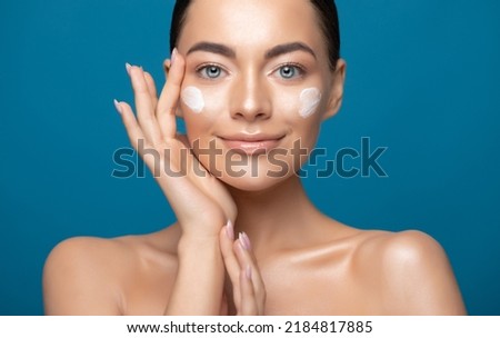 Beautiful young woman applying cosmetic cream treatment on her face isolated on blue color background
