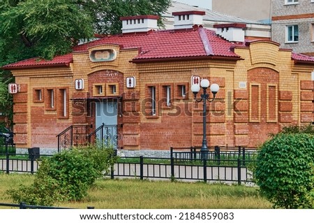 The building of a public toilet made of red brick on the city embankment. Green bushes and trees. Lanterns and fences of sidewalks. City infrastructure concept.