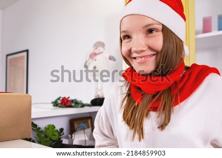 Blonde young woman sitting on her work place in the office in Santa hat and red scarf smiling, holiday concept and copy space.