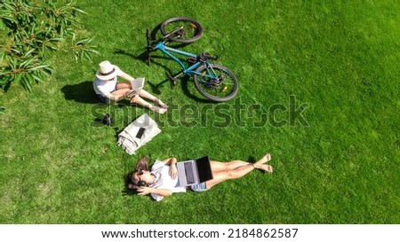 Young girls with bicycle in park, using laptop computer and headphones, two student girls studying online outdoors sitting on grass near bike, aerial drone view from above Royalty-Free Stock Photo #2184862587
