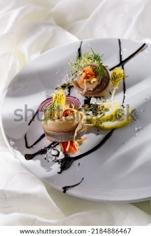 Baked mackerel with lemon slices, flower petals, parsley, onion and sauce on a white plate and light background. Vertical orientation