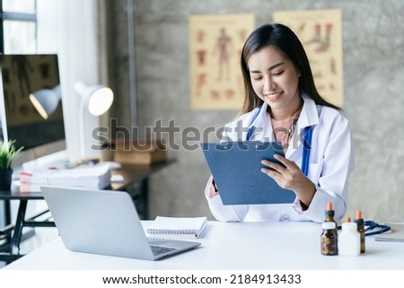 Smiling young Asian female doctor working at the clinic reception, she is using a computer and writing medical reports.