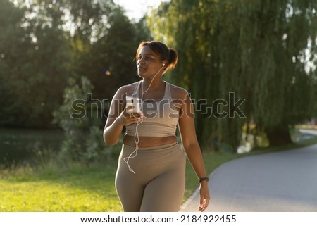 Plus sized African American woman using mobile phone and earphones during the walking at the park in a summer day  Royalty-Free Stock Photo #2184922455