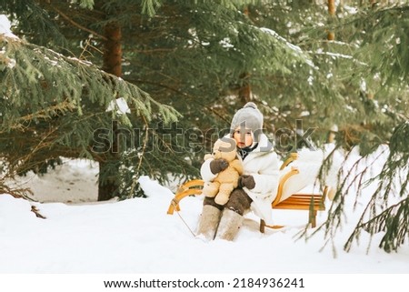 cute little boy toddler in fur coat, knitted hat and felt boots sitting on sled with teddy bear toy in snow winter forest, concept of Christmas holidays and New Year in retro vintage style