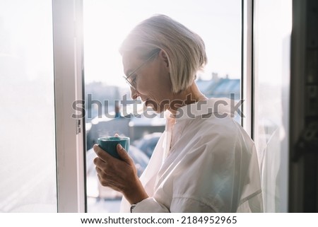Mature caucasian woman drinking tea or coffee from cup near window with view on blur city. Concept of domestic lifestyle. Partial image of modern successful lady wearing shirt and glasses. Sunny day