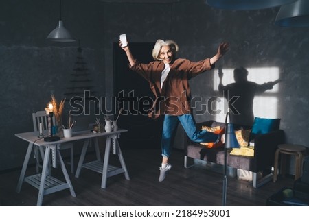 Pleased caucasian businesswoman with smartphone jumping at dark art studio with lighting. Small business and entrepreneurship. Modern successful woman rejoicing win and looking at camera