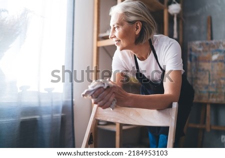 Smiling caucasian 50 years old female sculptor cleaning hands with rag after making clay. Small business and entrepreneurship. Home hobby, entertainment and leisure. Woman looking away. Art studio