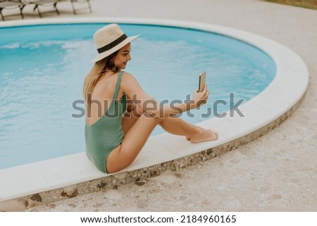 Cute young woman sitting by the swimming pool and taking selfie photo with  mobile phone in the house backyard