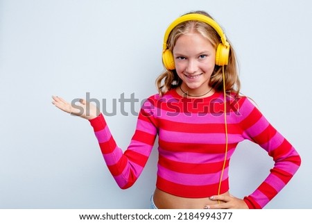 Caucasian teen girl listening to music isolated on blue background showing a copy space on a palm and holding another hand on waist.