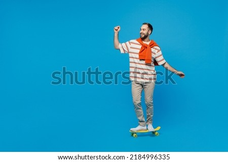 Full body side view young smiling happy fun cool caucasian man 20s wearing orange striped t-shirt riding yellow skateboard isolated on plain blue background studio portrait. People lifestyle concept