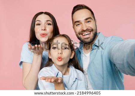 Close up young happy lovely parents mom dad with child kid daughter teen girl in blue clothes doing selfie shot pov on mobile cell phone blow air kiss isolated on plain pastel light pink background.