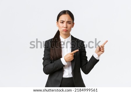 Disappointed or jealous female asian entrepreneur in suit complaining, pouting and pointing fingers right. Displeased saleswoman arguing, look offended or sad, standing white background