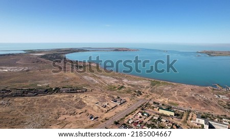 Drone view of Bertys Bay, Lake Balkhash. The blue-green water of the lake reflects the sky. There is a town and a factory on the shore. The coastal line is clearly visible, low houses