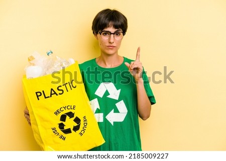 Young caucasian woman holding a bag full of plastic bottles to recycle isolated on yellow background showing number one with finger.